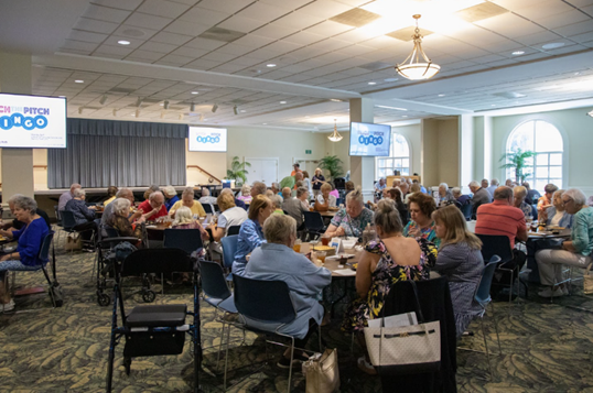 A group of people sit at various tables while watching a presentation.